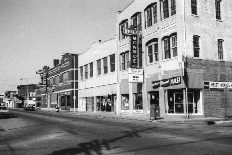 Black and white photo of a vintage street scene with old buildings and signs, clear sky, and a parked car.