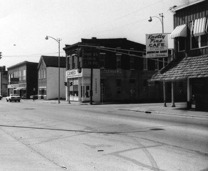 Black and white photo of a town street with buildings, a cafe, a cameo sign, and sparse traffic.