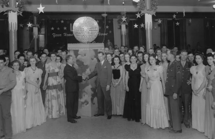 A large group of elegantly dressed people at a formal event, with a disco ball and festive decorations in the background.