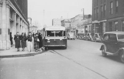 Vintage street scene with a trolley bus, old cars, and people waiting, suggesting it's from the early-mid 20th century.