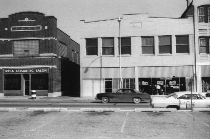 A black and white photo of a street scene with a cosmetic salon, cars parked, and other buildings.
