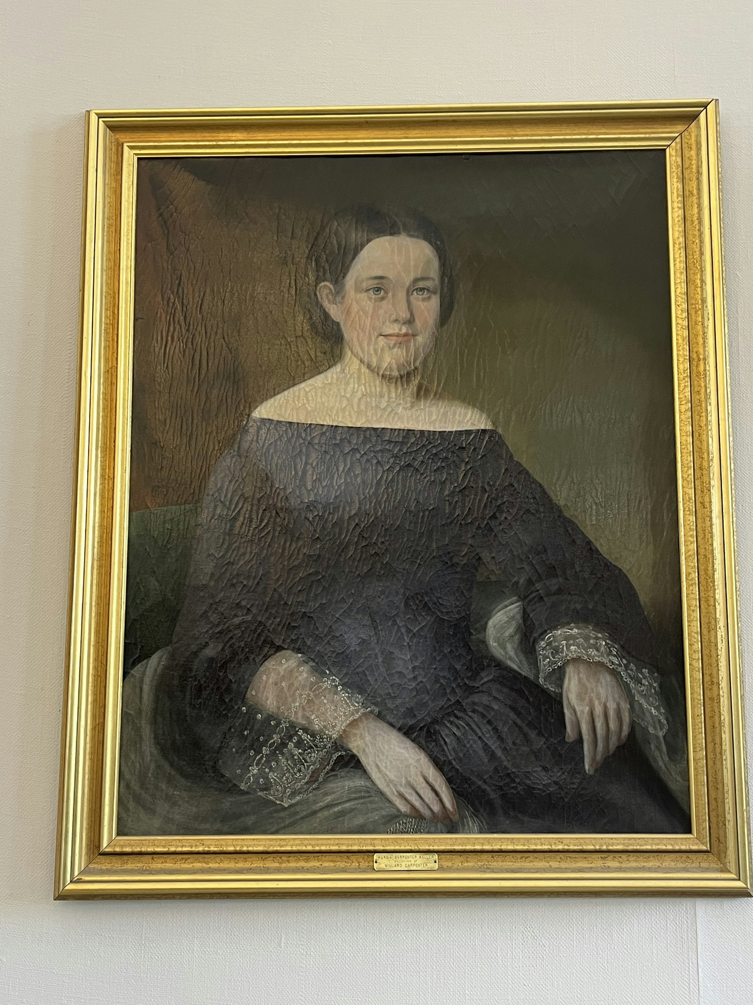 A traditional portrait of a woman in a dark dress with lace details, displayed in a gold frame.