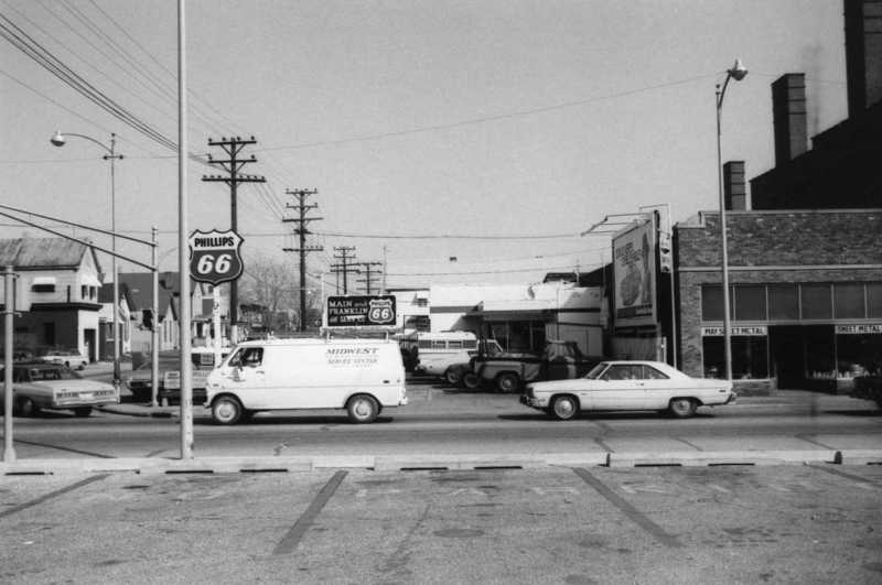 Black and white street view with vintage cars, gas station sign, and Route 66 marker.