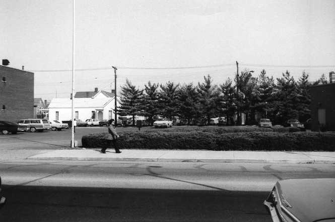 Black and white photo of a street, person walking, cars, trees, and buildings. Vintage vibe.
