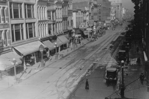 Vintage street scene with old buildings, cars, tram tracks, and a pedestrian.