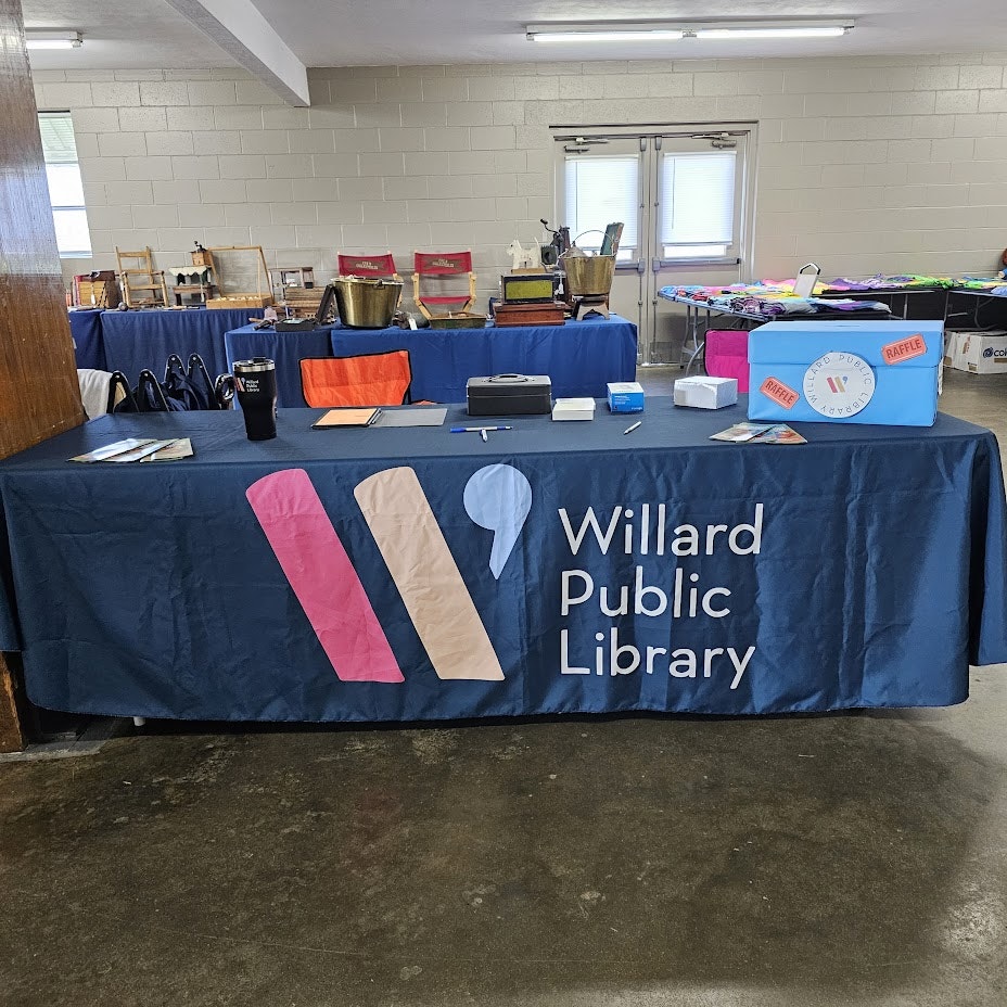 A table with a "Willard Public Library" banner, items for a sale or event, and a raffle wheel.