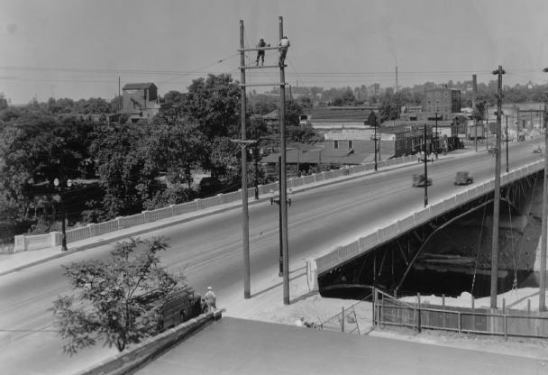 Black and white photo of an old street with cars, an overpass, utility poles, and buildings in the background.
