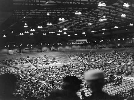 Indoor arena with audience seating and a stage; event in progress, captured in black and white.