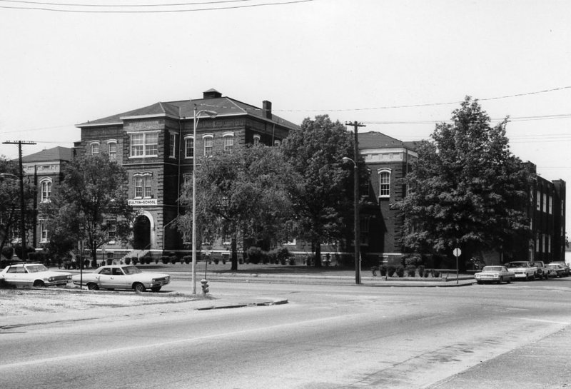 A vintage black and white photo showing an old building, trees, and parked cars on a sunny day.