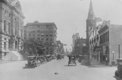Historic street scene with old cars, buildings, and a tram track.