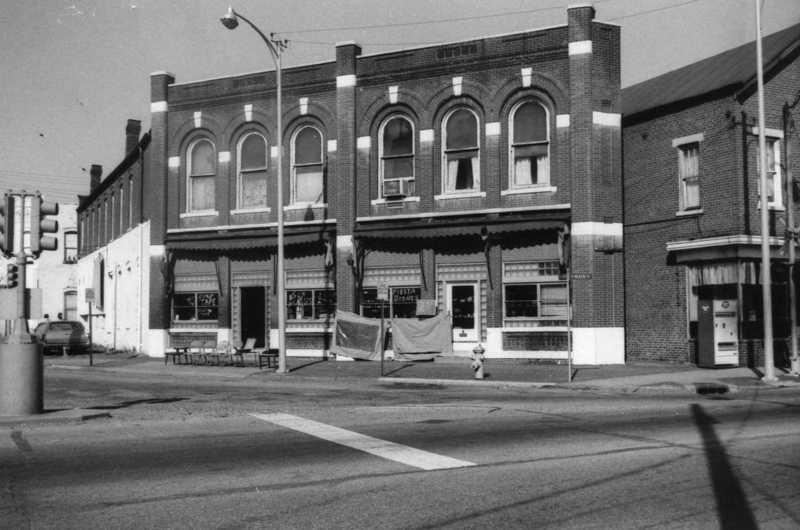 Black and white photo of an old-fashioned street corner with vintage-looking buildings and awnings.