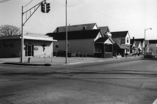 A black and white photo of a street corner showing buildings, a traffic light, and clear skies.