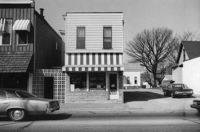 Vintage black and white photo of a small town street with an old car and storefront.