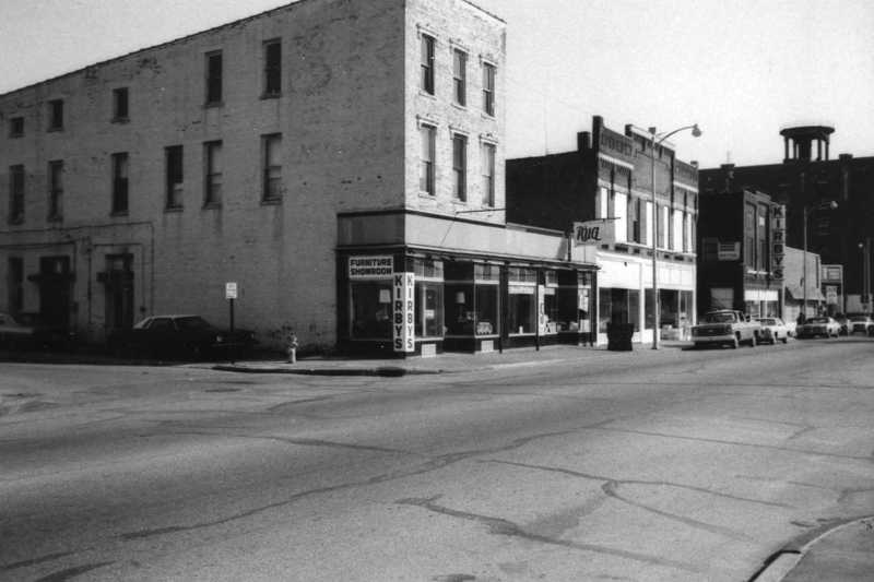 Black and white photo of a vintage street corner with old buildings and parked cars.