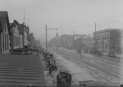 A black and white photo of an early 20th-century street scene with trams, vintage cars, and buildings.