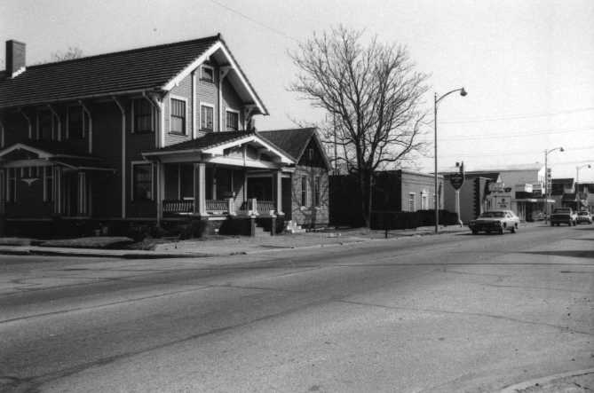 Black and white photo of a residential street with houses, clear sky, and a leafless tree.