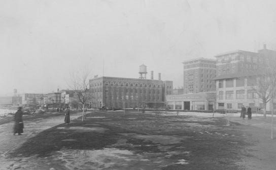 Black & white photo of an old city square with vintage buildings and few pedestrians, possibly early 20th century.