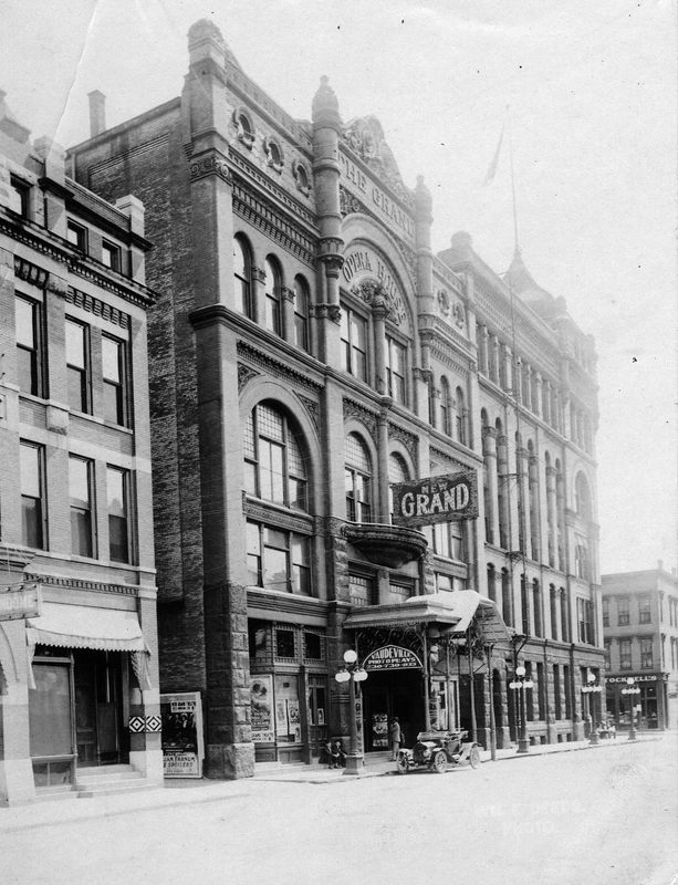 Historic photo of a theater named "Grand" with ornate architecture, advertising Vaudeville, and an early car parked outside.