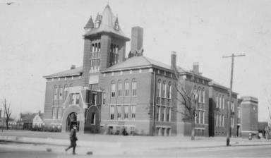 Vintage photo of an old building with a tower, a person walking by, and a street in the foreground.