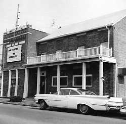 Vintage car parked outside an old building with balcony and antennas. Black and white photo.