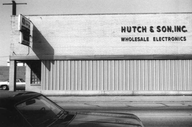 A black-and-white photo of a building with a sign "HUTCH & SON, INC. WHOLESALE ELECTRONICS" and part of a car in the foreground.