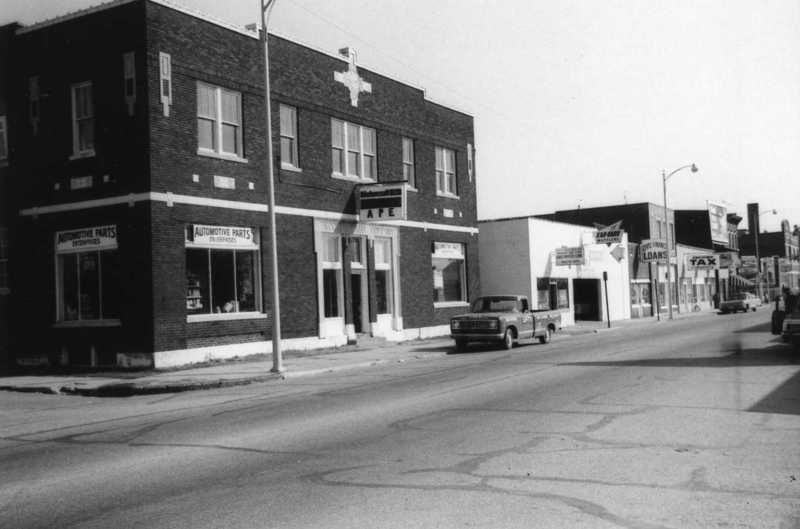 Vintage street scene with buildings, a cafe, automotive parts shop, cars, and other businesses.