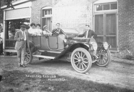 Vintage photo of people with an early 1900s car, outside a brick building, possibly a garden or park entrance.