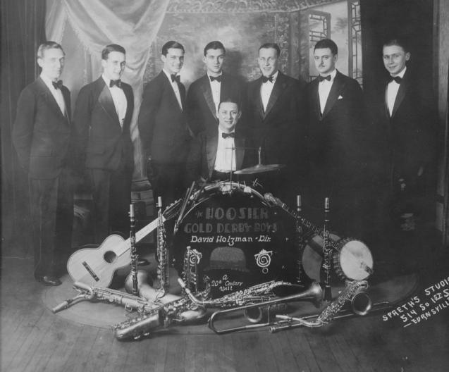 Seven men in tuxedos with a drum kit and various instruments, likely a band.