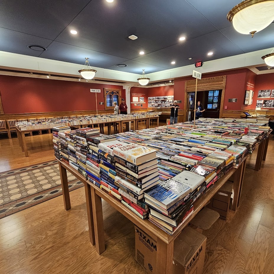 A book sale in a room with tables filled with organized books and people browsing in the background.