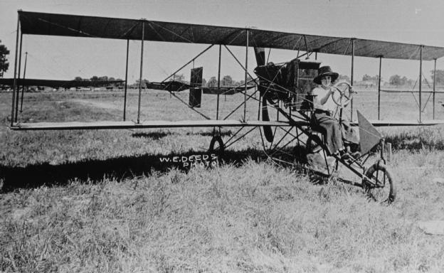 A historical black and white photo of an early biplane with a pilot and an observer standing nearby in a grassy field.