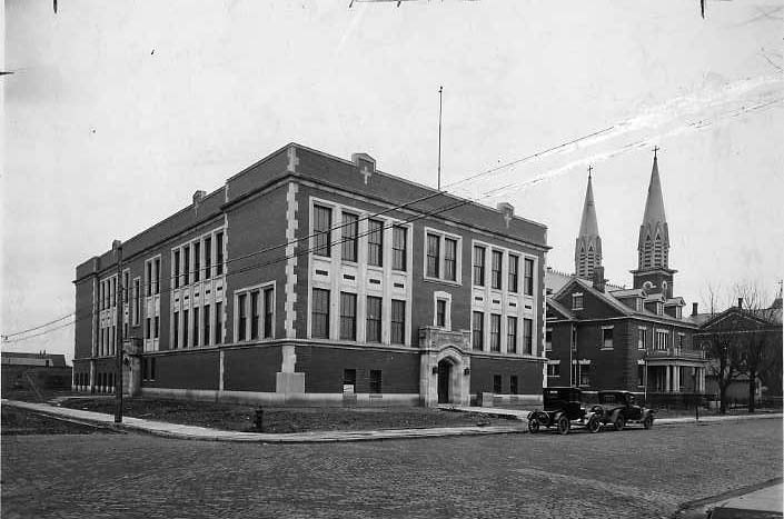 A vintage photo: classic building, church spires in the back, old cars, cobblestone street.