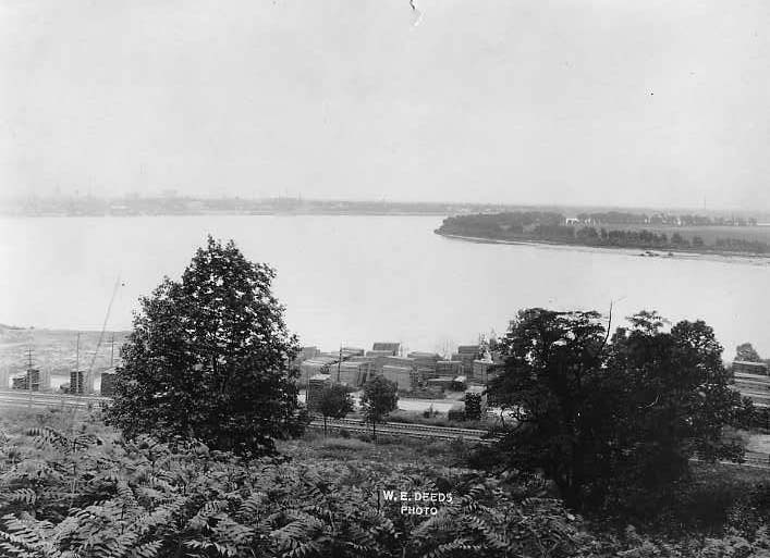 Black and white photo of a river bend, buildings along the shore, and vegetation in the foreground.