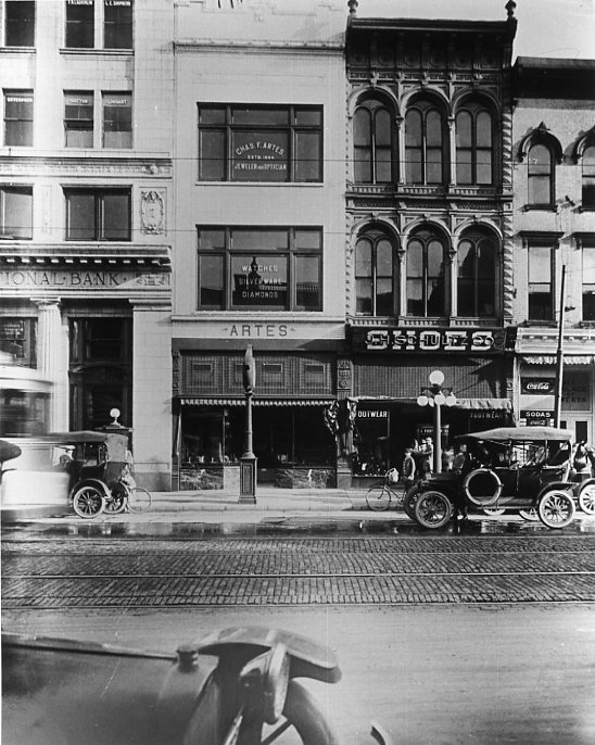 Vintage street scene with old buildings, cars, and shops, such as a bank and jewelers.