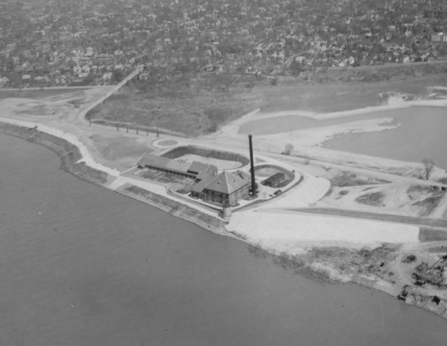 Aerial view of a coastal industrial facility with a tall chimney, near a residential area.