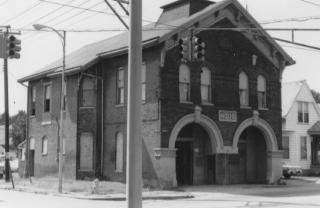 Old two-story building on a street corner with traffic lights and power lines. Black and white photo.