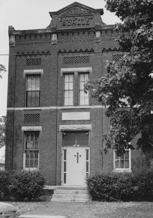 Old brick building with "EVANSVILLE SCHOOL" sign, tall windows, and a doorway. Vintage aesthetic. Vehicle partial view.