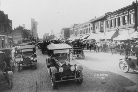 Black and white photo of an early 20th-century street scene with vintage cars and horse-drawn carriages.