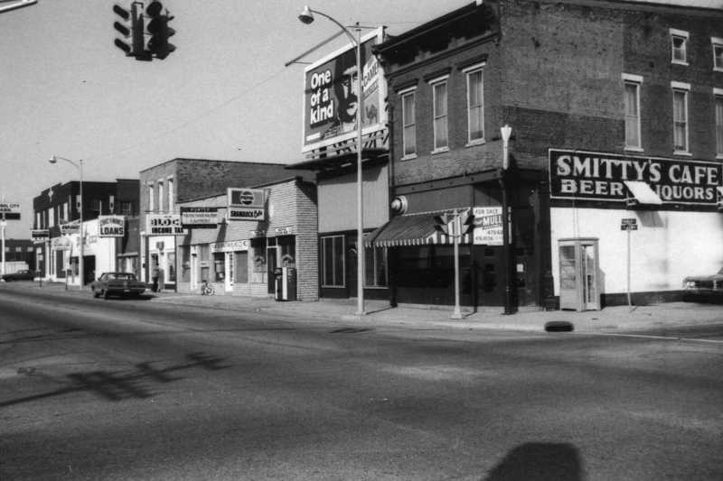 A vintage street view with shops, a "one of a kind" sign, and sparse traffic. Black and white photo.