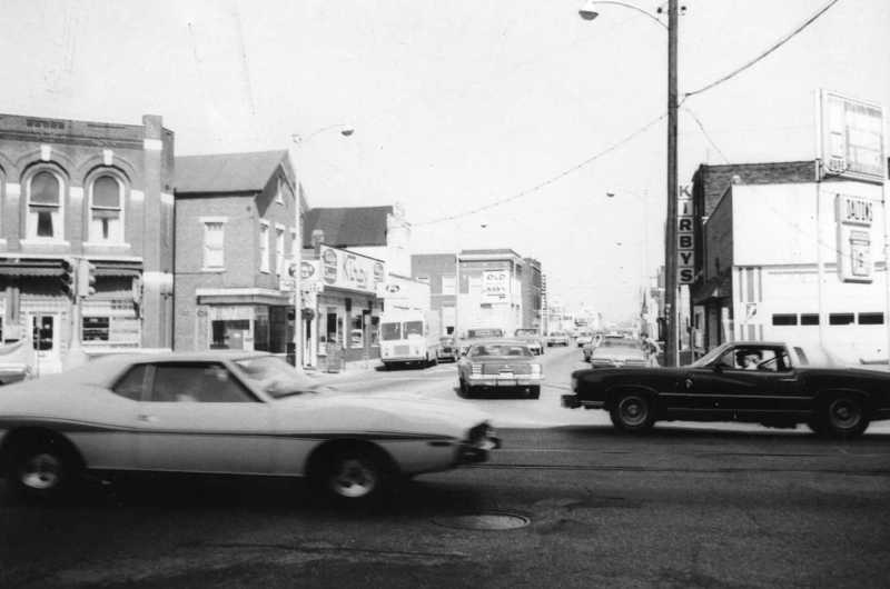 Vintage black-and-white photo of a street with old cars and classic storefronts.
