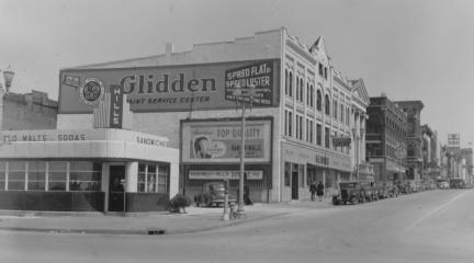 Vintage street view with old buildings, signs for businesses like "Glidden" and cars parked along the curb, appearing historical.