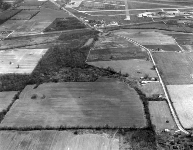 Aerial black and white photo showing farmland with fields, roads, and a few buildings.