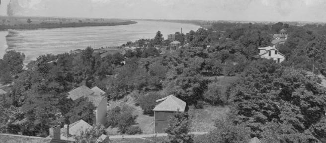 A historic black and white photo of a riverside settlement with buildings amidst abundant trees.