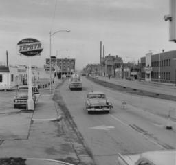 Vintage street scene with cars, signs, and buildings, in black and white.