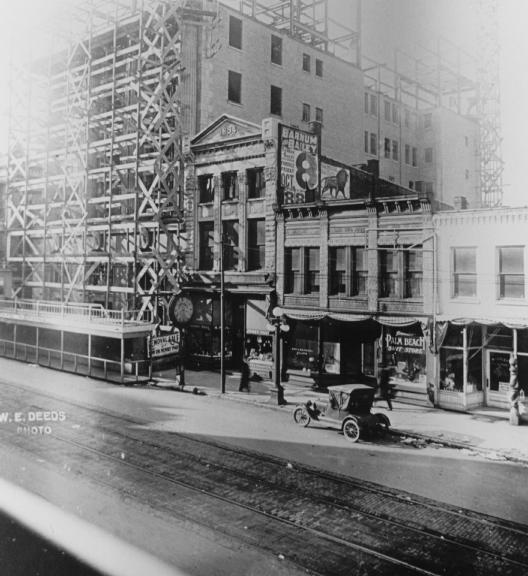 Vintage street scene with buildings, scaffolding, an old car, and tram tracks.