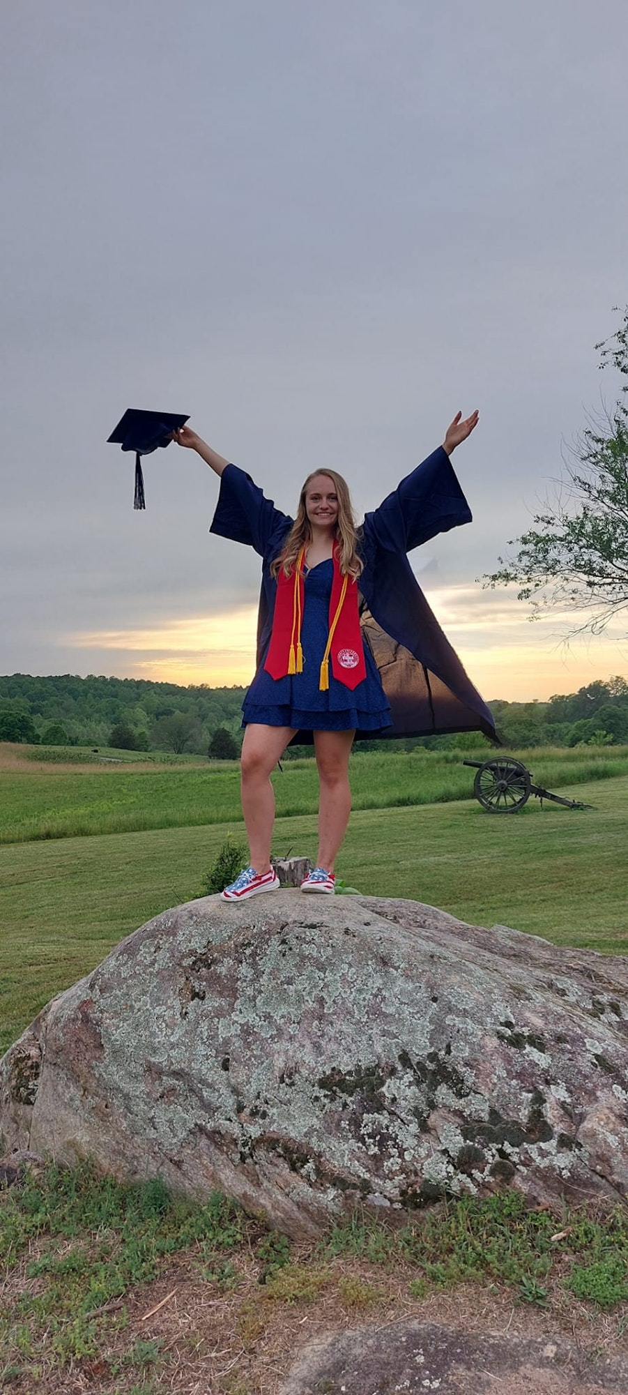 A graduate in a cap and gown stands on a rock, arms raised, with a cannon and scenic landscape in the background.