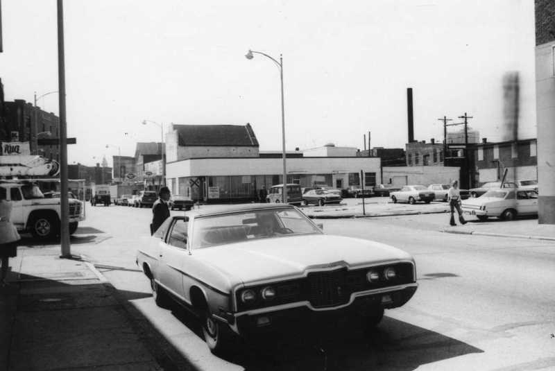 Black and white photo of a vintage car parked on a street; streetlights and buildings in the background.