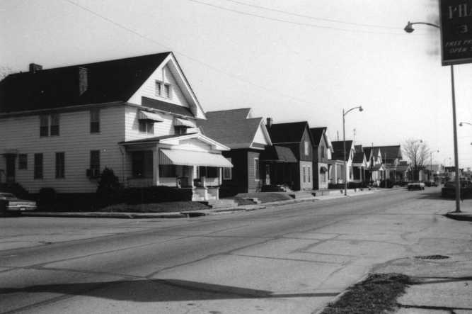 A black and white photo of a suburban street with residential houses and a street sign.