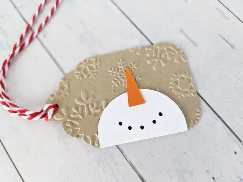 A craft paper tag with a snowman design and embossed snowflakes, attached to a red and white string.