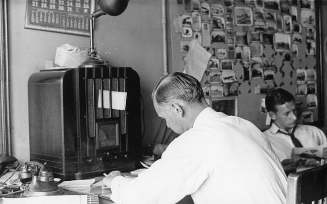 Two men at a desk, one writing, with a vintage radio and wall filled with photos.