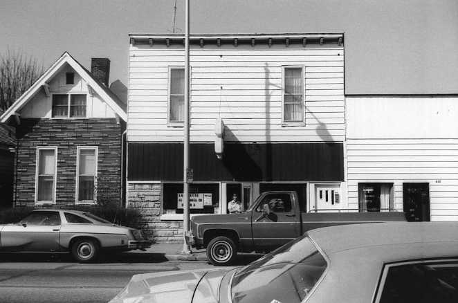 Black and white photo of a street with cars and buildings, including a house with a storefront.